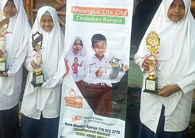 Santriwati Pondok Roja memegang piala prestasi di samping banner BMH Merangkai Cita-Cita Cerdaskan Bangsa