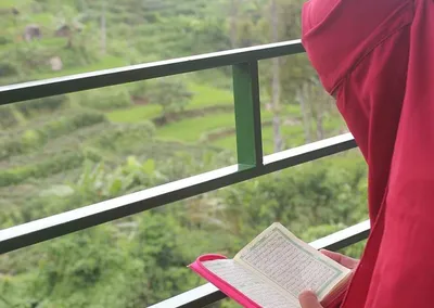Santriwati berkerudung merah membaca mushaf Al-Qur’an kecil di balkon menghadap perbukitan hijau