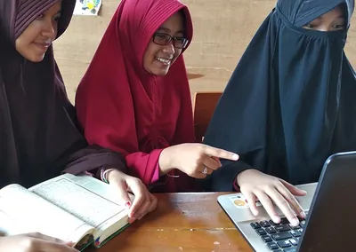 Tiga santriwati belajar bersama dengan mushaf Al-Qur’an dan laptop di ruang kelas